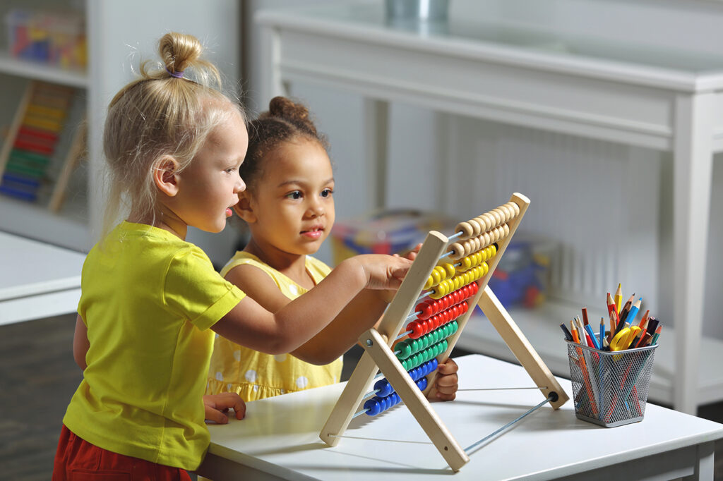 Two small children standing at a table move their fingers along an abacus on the table.